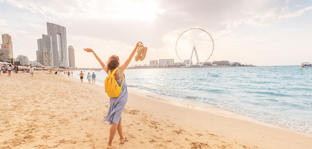 Happy female tourist walks barefoot on a sandy beach in the JBR area of Dubai and admires the panoramic view of the Ain Ferris Wheel